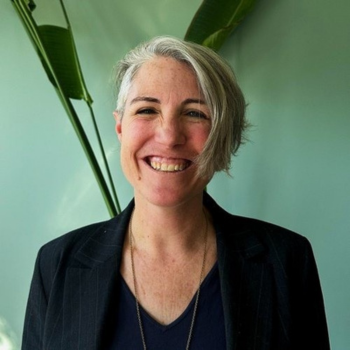 A person smiling warmly in front of a light green wall with large leaves behind them.